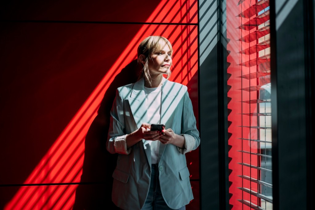 Nachdenkliche blonde Geschäftsfrau mit Smartphone schaut durch Fenster im Büro.