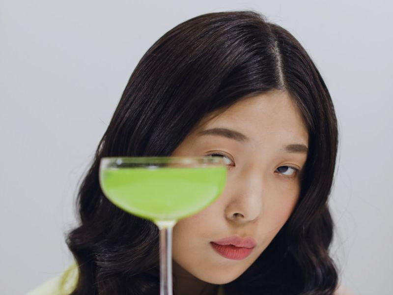 Frau schaut hinter einem mit grüner Flüssigkeit gefülltem Glas hervor.