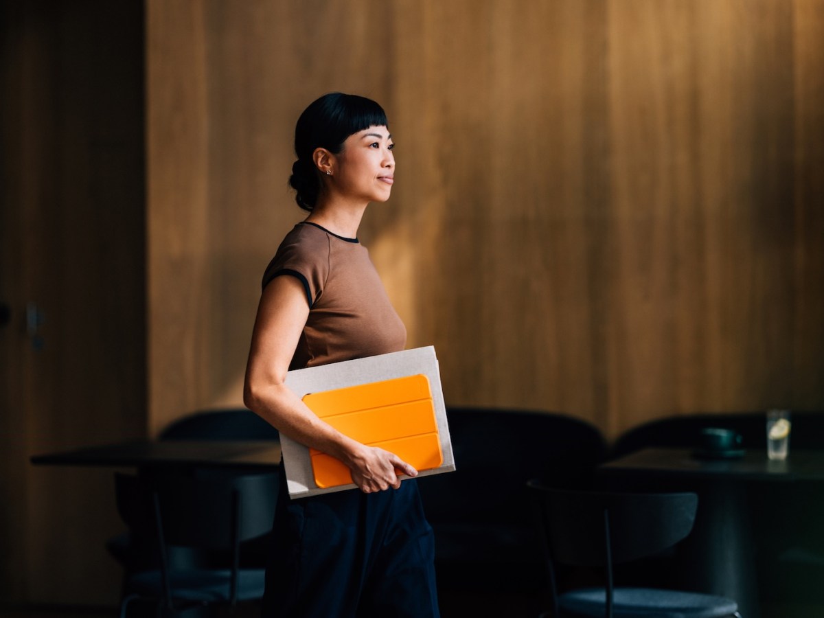 Eine Frau betritt mit einem leuchtend orangefarbenen Notizbuch in der Hand ein modernes Büro.