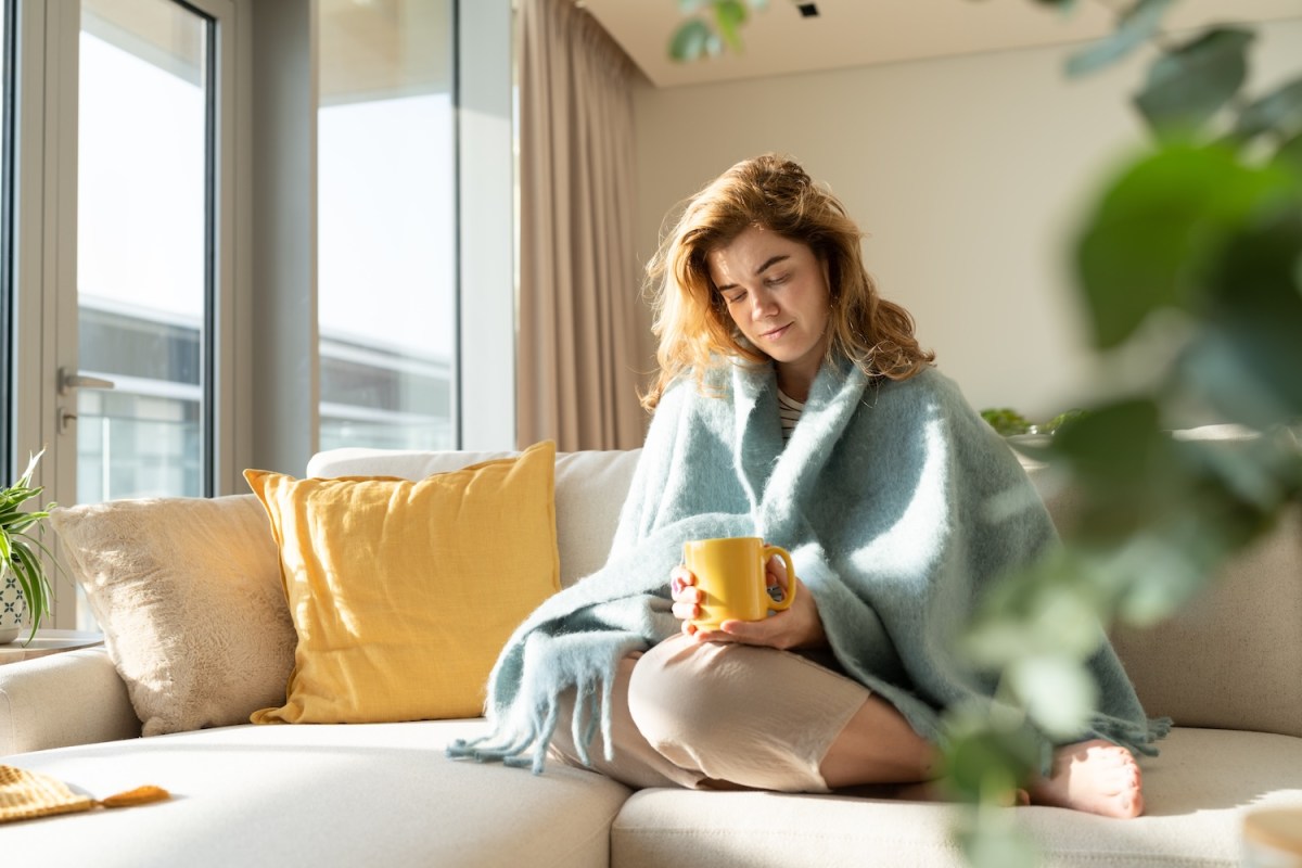 Junge Frau in Decke gehüllt macht eine Pause mit einer Tasse Tee im Wohnzimmer.