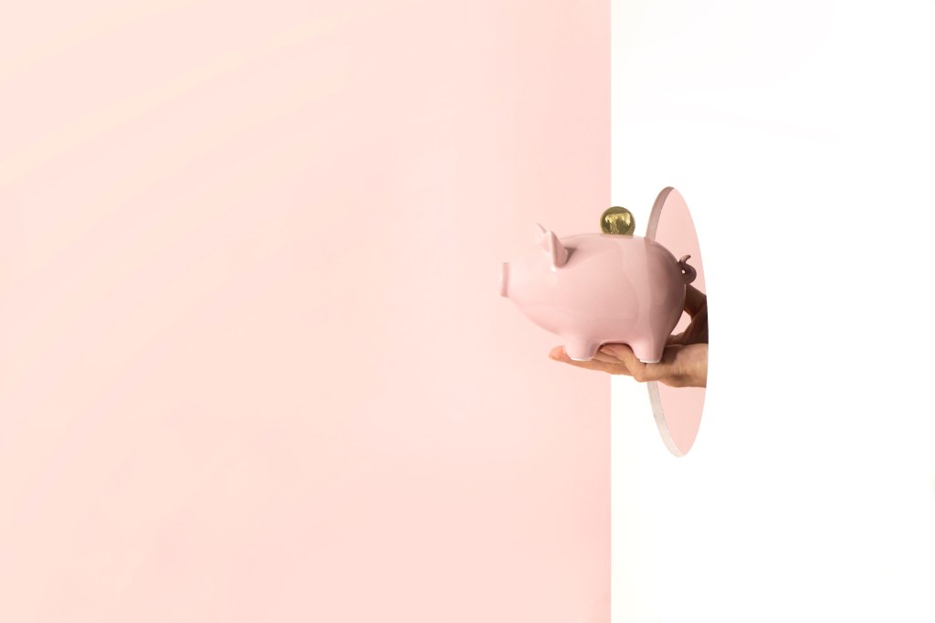 Eine Hand hält ein Sparschwein durch eine Öffnung an einer weißen Wand.