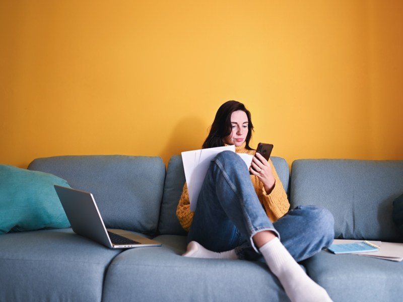 Junge Frau sitzt auf dem Sofa und schaut auf ihr Handy und hat Unterlagen in der Hand.