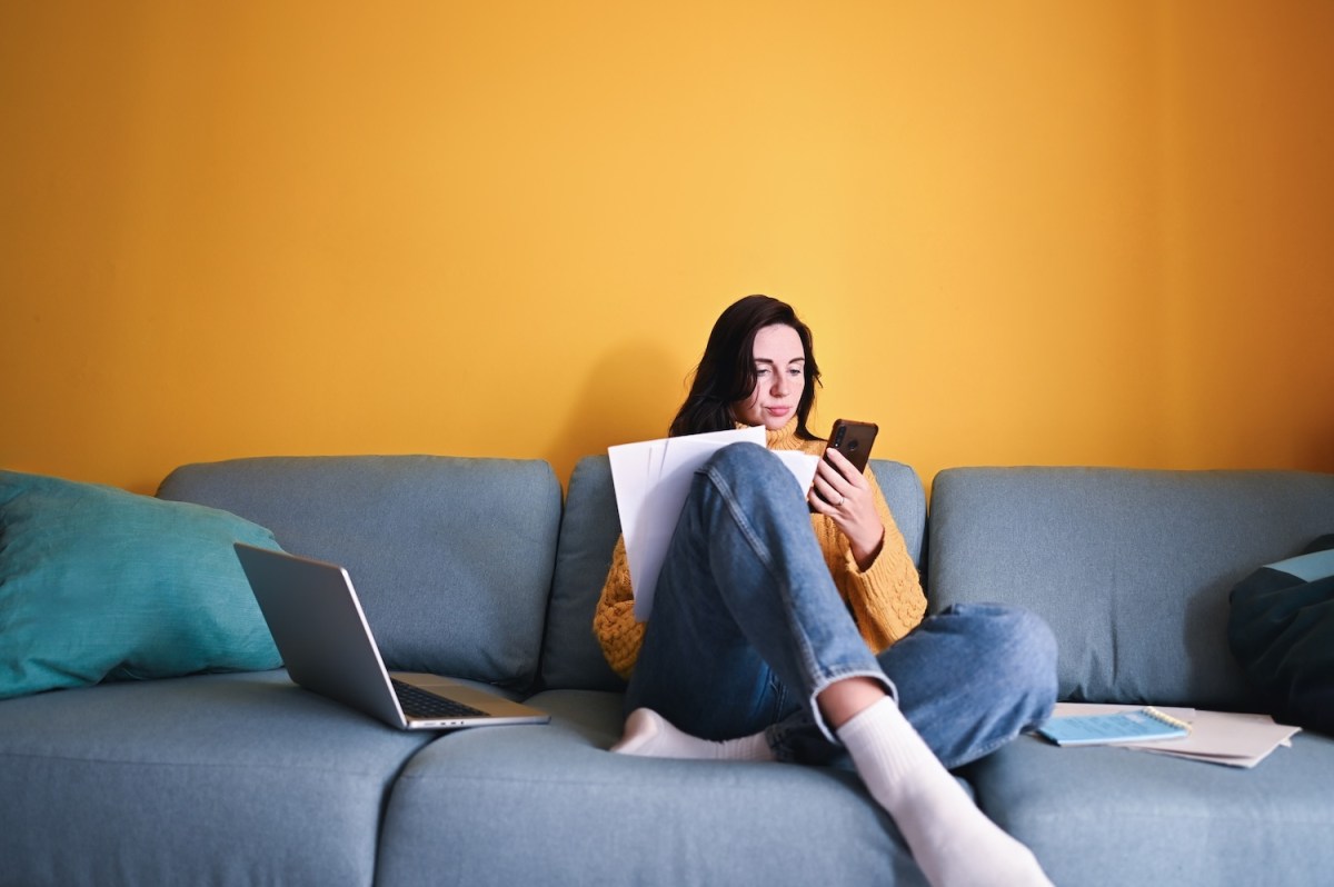 Junge Frau sitzt auf dem Sofa und schaut auf ihr Handy und hat Unterlagen in der Hand.