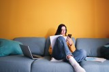 Junge Frau sitzt auf dem Sofa und schaut auf ihr Handy und hat Unterlagen in der Hand.