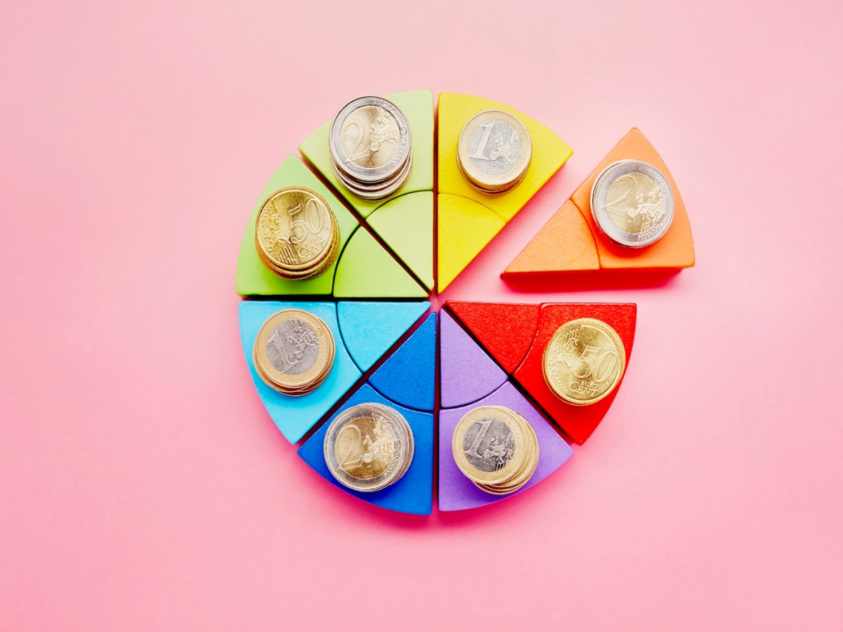 Kreisdiagramm aus bunten Bausteinen und Stapeln von Euro-Münzen auf rosa Hintergrund.