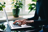 Frau tippt auf einem Laptop an einem Schreibtisch mit Fenstern und grünen Pflanzen.