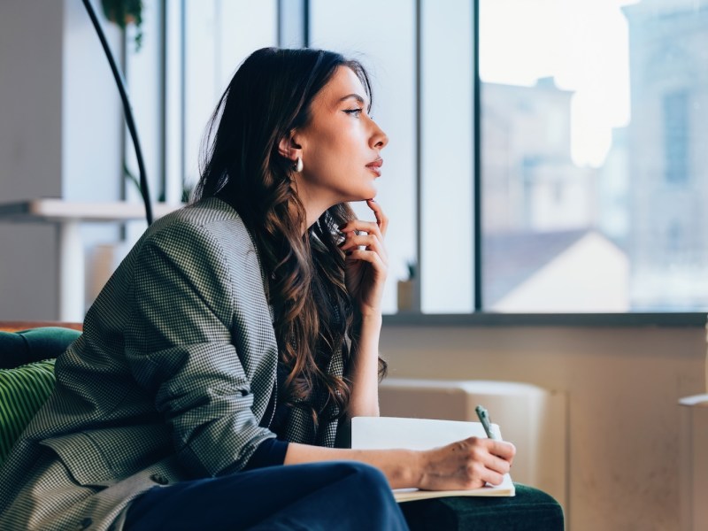 Eine berufstätige Frau sitzt nachdenklich mit Notizbuch und Stift in der Hand in einer modernen Büroumgebung mit großen Fenstern.