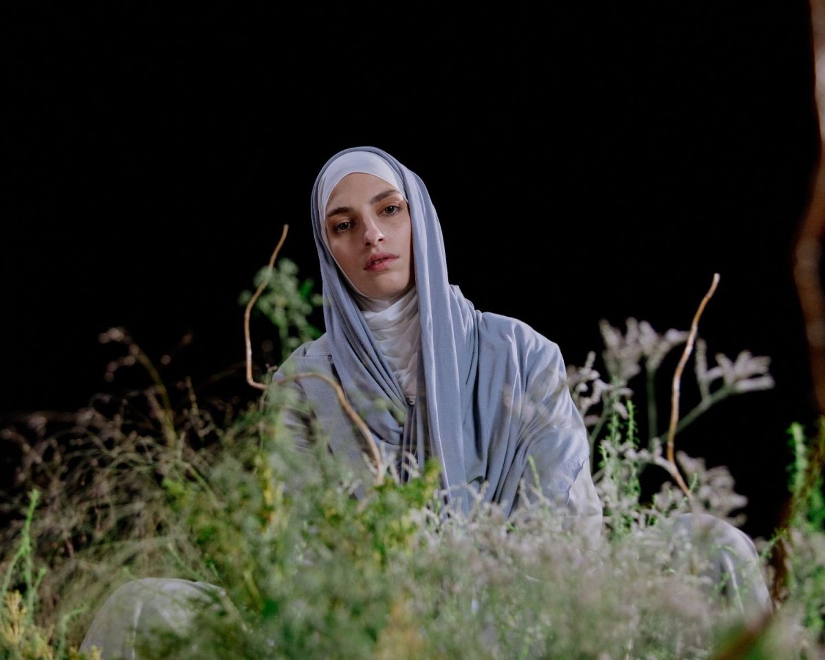 Frau mit einem Kopftuch steht hinter einem Strauch vor einem schwarzen Hintergrund.