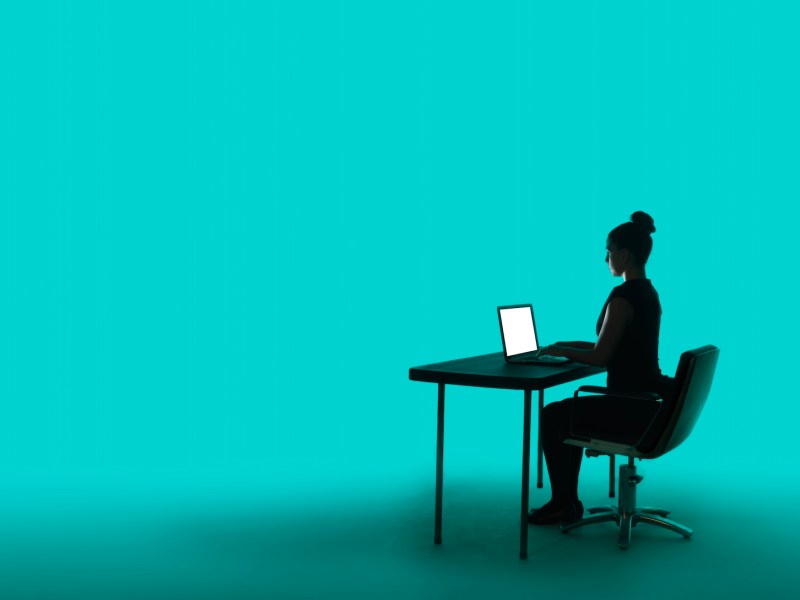 Junge Geschäftsfrau in Silhouette, sitzt an ihrem Schreibtisch mit beleuchtetem Laptop, auf kräftigem türkisfarbenem Hintergrund.