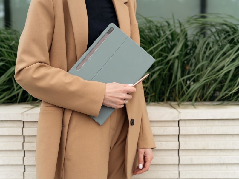 Eine Frau in beigen Blazer hält einen Laptop in der Hand.