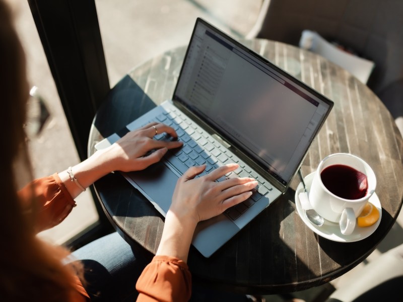 Freiberuflerin arbeitet am Laptop und trinkt Tee an einem Fenstertisch.