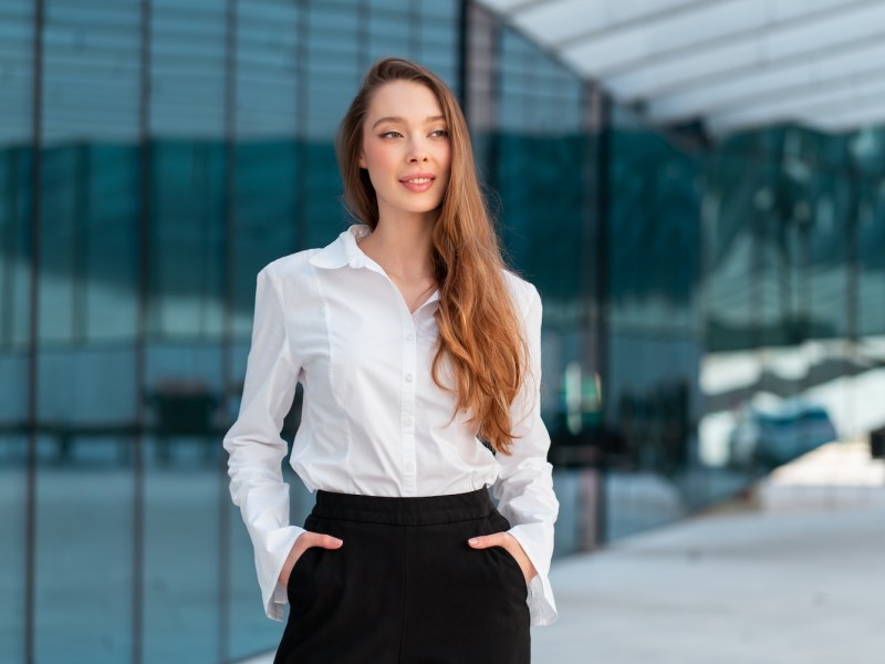 Selbstbewusste Geschäftsfrau mit langen Haaren steht vor einem modernen Bürogebäude.