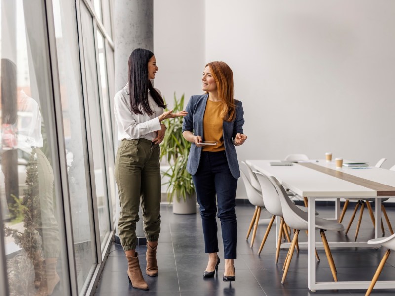 Zwei Geschäftsfrauen in eleganter Freizeitkleidung gehen in der Firmenzentrale spazieren und führen ein Gespräch.