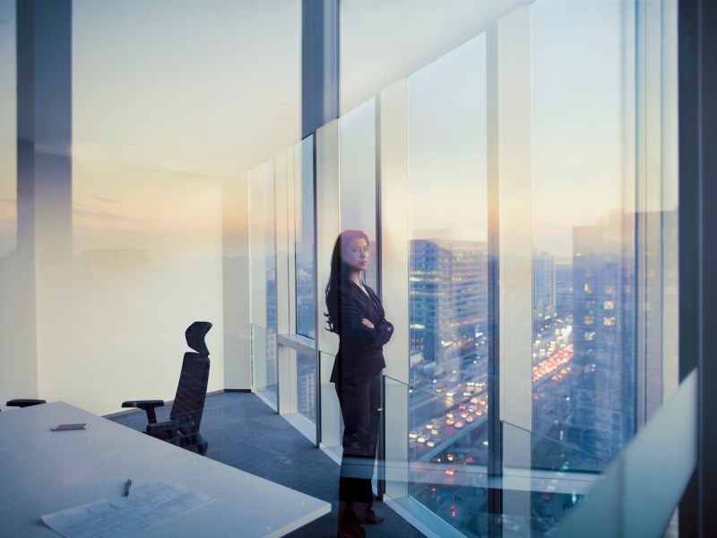 Eine Frau im Anzug schaut nachdenklich aus dem Fenster eines Meetingraums.
